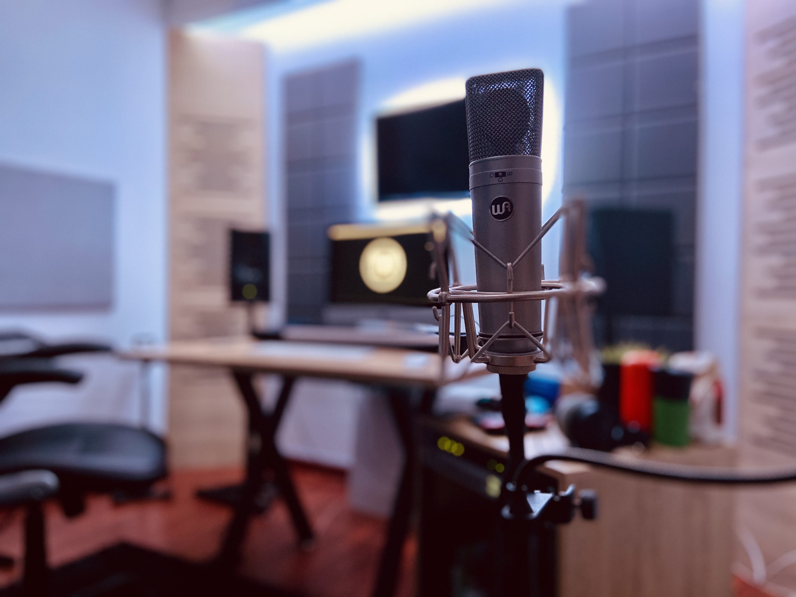 A condenser microphone in a home studio setup with a computer and studio monitors in the background.
